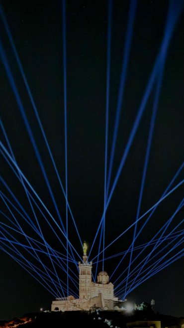 <!-- wp:heading {"level":1} -->
<h1 class="wp-block-heading">Ce dimanche 7 décembre 2025, Marseille a vibré au rythme d’un événement exceptionnel : la (re)découverte de la statue rénovée de Notre-Dame de la Garde — la « Bonne-Mère » — sublimée, pour l’occasion, par un spectaculaire show son et lumière. Un moment de fête, d’émotion et de communion, qui a offert à la cité phocéenne un regard renouvelé sur son monument emblématique.</h1>
<!-- /wp:heading -->

<!-- wp:heading -->
<h2 class="wp-block-heading">Un chantier titanesque pour redonner vie à un symbole</h2>
<!-- /wp:heading -->

<!-- wp:paragraph -->
<p>La statue de la Vierge, perchée au sommet de la basilique à quelque 210-220 m d’altitude, avait besoin d’un coup de jeune : les outrages du temps, le sel marin, le mistral et la pollution avaient terni l’éclat d’origine.&nbsp;</p>
<!-- /wp:paragraph -->

<!-- wp:paragraph -->
<p>Début 2025, un vaste chantier a été lancé par le Diocèse de Marseille, alliant savoir-faire traditionnel et techniques modernes.&nbsp; Pas moins de 30 000 à 40 000 feuilles d’or ont été appliquées à la main sur la statue, recouvrant plus de 100 m² de surface : une dorure pure, minutieuse, qui redonne à la « Bonne-Mère » tout son lustre.&nbsp;</p>
<!-- /wp:paragraph -->

<!-- wp:paragraph -->
<p>Mais ce n’est pas tout : la restauration n’a pas concerné que la dorure. La structure interne de la statue a été traitée contre la corrosion, le piédestal, les pierres du clocher, les gargouilles et les éléments architecturaux de la basilique ont eux aussi été remis en état.&nbsp;</p>
<!-- /wp:paragraph -->

<!-- wp:paragraph -->
<p>Ce chantier monumental, d’un coût total estimé à environ 2,8 millions d’euros, a été rendu possible grâce à des dons, le soutien de collectivités et de mécènes.&nbsp;</p>
<!-- /wp:paragraph -->

<!-- wp:heading -->
<h2 class="wp-block-heading">Une cérémonie, une procession, une célébration</h2>
<!-- /wp:heading -->

<!-- wp:paragraph -->
<p>La journée du 7 décembre a débuté dans un cadre spirituel et solennel. À 15h30, à l’Abbaye Saint-Victor, une messe d’action de grâce a rassemblé fidèles et habitants.&nbsp;</p>
<!-- /wp:paragraph -->

<!-- wp:paragraph -->
<p>Puis, à 16h30, le public a été convié à se rassembler sur la place Joseph-Étienne pour le départ d’une procession — un moment de recueillement et de partage — qui a mené jusqu’à la colline de <a href="https://basiliquenotredamedelagarde.com" target="_blank" rel="noopener" title="Notre-Dame de la Garde.">Notre-Dame de la Garde.</a> </p>
<!-- /wp:paragraph -->

<!-- wp:paragraph -->
<p>Le point d’orgue a été marqué à 18h, avec une action de grâce et la bénédiction de la statue rénovée, en présence de Jean‑Marc Aveline, archevêque de Marseille.&nbsp;</p>
<!-- /wp:paragraph -->

<!-- wp:heading -->
<h2 class="wp-block-heading">Quand la lumière sublime le patrimoine : le spectacle « Élévation »</h2>
<!-- /wp:heading -->

<!-- wp:paragraph -->
<p>Dès la tombée de la nuit, la basilique et la colline se sont transformées en écrin pour un spectacle éblouissant. Conçu par le collectif Monumental Tour et orchestré par le DJ français Michaël Canitrot, le show son et lumière intitulé « Élévation » a mêlé projections lumineuses, musique électronique et orchestrale, chants lyriques et chorale.&nbsp;</p>
<!-- /wp:paragraph -->

<!-- wp:paragraph -->
<p>Pendant environ douze minutes, quatre tableaux se sont succédé pour raconter l’histoire de la Bonne-Mère :</p>
<!-- /wp:paragraph -->

<!-- wp:list -->
<ul class="wp-block-list"><!-- wp:list-item -->
<li>le lever du soleil, symbole de renouveau et d’espoir,</li>
<!-- /wp:list-item -->

<!-- wp:list-item -->
<li>le lien avec la mer et les marins, rappelant l’histoire maritime de Marseille,</li>
<!-- /wp:list-item -->

<!-- wp:list-item -->
<li>l’élévation, entre ciel et terre, matière et spiritualité,</li>
<!-- /wp:list-item -->

<!-- wp:list-item -->
<li>et enfin un hymne à la restauration, à ceux qui ont œuvré pour redonner à la statue sa splendeur.  </li>
<!-- /wp:list-item --></ul>
<!-- /wp:list -->

<!-- wp:paragraph -->
<p>La mise en lumière a dévoilé la statue sous un jour nouveau, jouant sur les contrastes, les reflets dorés, les teintes marines ou cuivrées — un spectacle visuel et sonore, spectaculaire, émouvant, pensé comme un hommage à l’histoire de Marseille.&nbsp;</p>
<!-- /wp:paragraph -->

<!-- wp:heading -->
<h2 class="wp-block-heading">Une ville unie dans l’émerveillement</h2>
<!-- /wp:heading -->

<!-- wp:paragraph -->
<p>Même si l’accès immédiat à la basilique était fermé au public pour des raisons de sécurité (le périmètre réservé aux organisateurs)&nbsp; , Marseille tout entière — les quartiers, les collines, les canaux visuels — est devenue une scène.</p>
<!-- /wp:paragraph -->

<!-- wp:paragraph -->
<p>Des points de vue stratégiques avaient été annoncés pour profiter du spectacle en toute sécurité : depuis l’Hôtel de Ville, les jardins du Palais du Pharo, <a href="https://hotelbellevuemarseille.com/hotel/" target="_blank" rel="noopener" title="L'Hôtel Belle-vue">L'Hôtel Belle-vue</a> ou encore des zones en contrebas de la colline. </p>
<!-- /wp:paragraph -->

<!-- wp:paragraph -->
<p>Pour beaucoup, ce moment était plus qu’un spectacle : c’était une fête du patrimoine, un geste collectif, un symbole d’attachement à la ville. Comme le rappelait l’archevêque, la statue — perchée dans le ciel de Marseille — « fait partie de la famille des Marseillais ».&nbsp;</p>
<!-- /wp:paragraph -->

<!-- wp:image {"id":17094,"sizeSlug":"large","linkDestination":"none"} -->
<figure class="wp-block-image size-large"><img src="https://hotelbellevuemarseille.com/wp-content/uploads/2025/12/BCFC2F78-4EBB-45FA-B63E-F44D971B59AF-576x1024.jpg" alt="inauguration Notre Dame de La Garde Marseille spectacle" class="wp-image-17094"/><figcaption class="wp-element-caption"></figcaption></figure>
<!-- /wp:image -->

<!-- wp:heading -->
<h2 class="wp-block-heading">Au-delà de la dorure : un symbole renouvelé</h2>
<!-- /wp:heading -->

<!-- wp:paragraph -->
<p>La restauration de la Bonne-Mère, et sa mise en lumière ce soir, dépasse largement l’aspect esthétique. C’est une reconquête du patrimoine, un acte de mémoire, un geste d’amour pour Marseille.</p>
<!-- /wp:paragraph -->

<!-- wp:paragraph -->
<p>C’est aussi le témoignage d’un engagement — celui d’artisans, de donateurs, d’institutions, de citoyens — pour préserver un héritage commun. Grâce à ce chantier, la statue retrouvera non seulement sa splendeur, mais aussi une protection durable contre les ravages du temps et des éléments.&nbsp;</p>
<!-- /wp:paragraph -->

<!-- wp:paragraph -->
<p>Le spectacle « Élévation » a montré que le patrimoine peut aussi se vivre, s’expérimenter, s’admirer ensemble — à travers l’art, la lumière, la musique — et devenir un moment de rassemblement, de fierté, de partage.</p>
<!-- /wp:paragraph -->

<!-- wp:paragraph -->
<p>La soirée du 7 décembre 2025 restera gravée dans la mémoire des Marseillais. La « Bonne-Mère », plus dorée que jamais, veille à nouveau sur la ville — mais aujourd’hui, elle brille, elle rayonne, elle rassemble.</p>
<!-- /wp:paragraph --> Hôtel écologique Belle-Vue vieux-port Marseille Dorure de la «Bonne-Mère», une soirée mémorable à Marseille